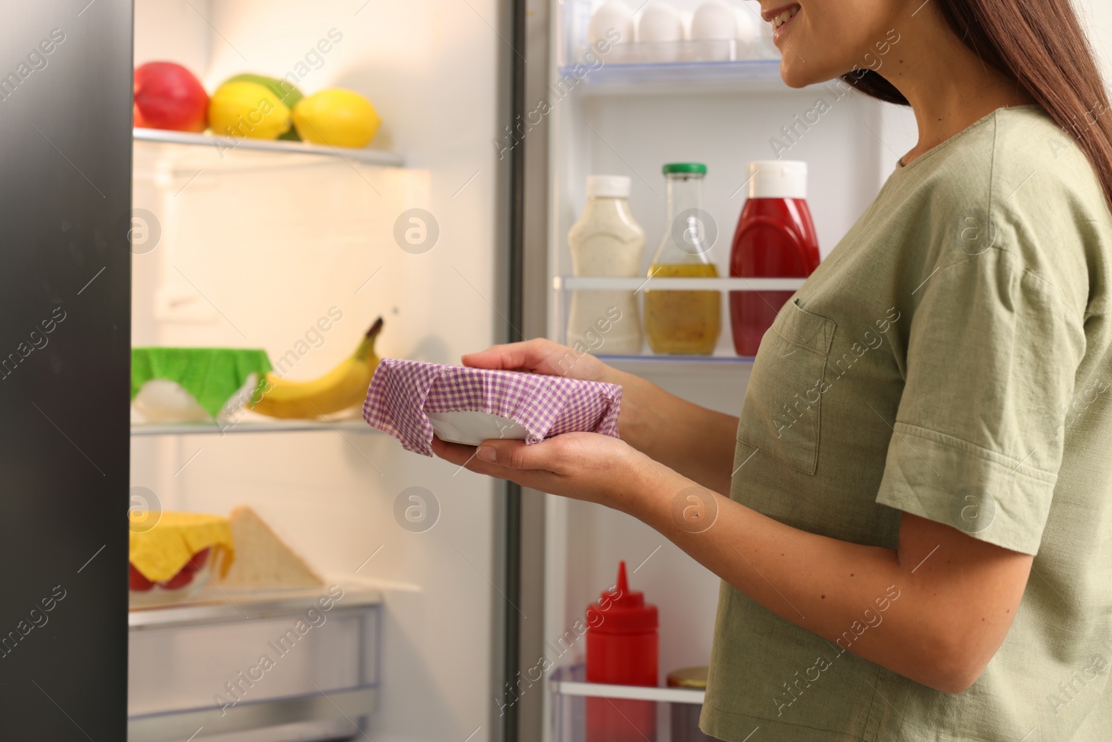 Photo of Woman holding bowl covered with beeswax food wrap near refrigerator in kitchen, closeup