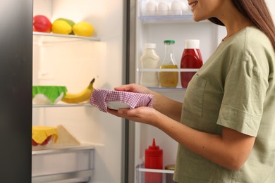 Woman holding bowl covered with beeswax food wrap near refrigerator in kitchen, closeup