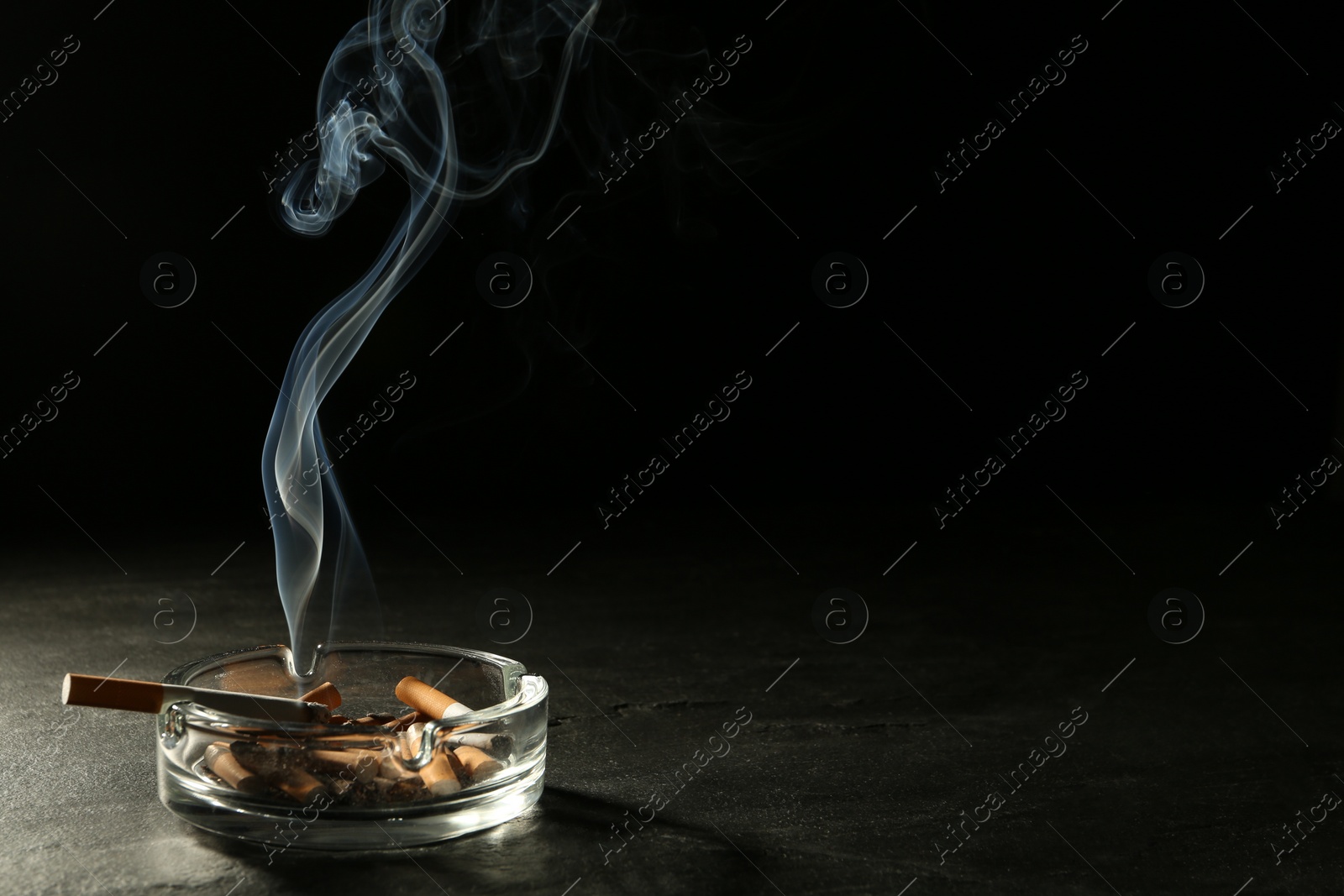 Photo of Smoldering cigarette in glass ashtray on grey table against black background. Space for text