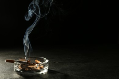 Photo of Smoldering cigarette in glass ashtray on grey table against black background. Space for text