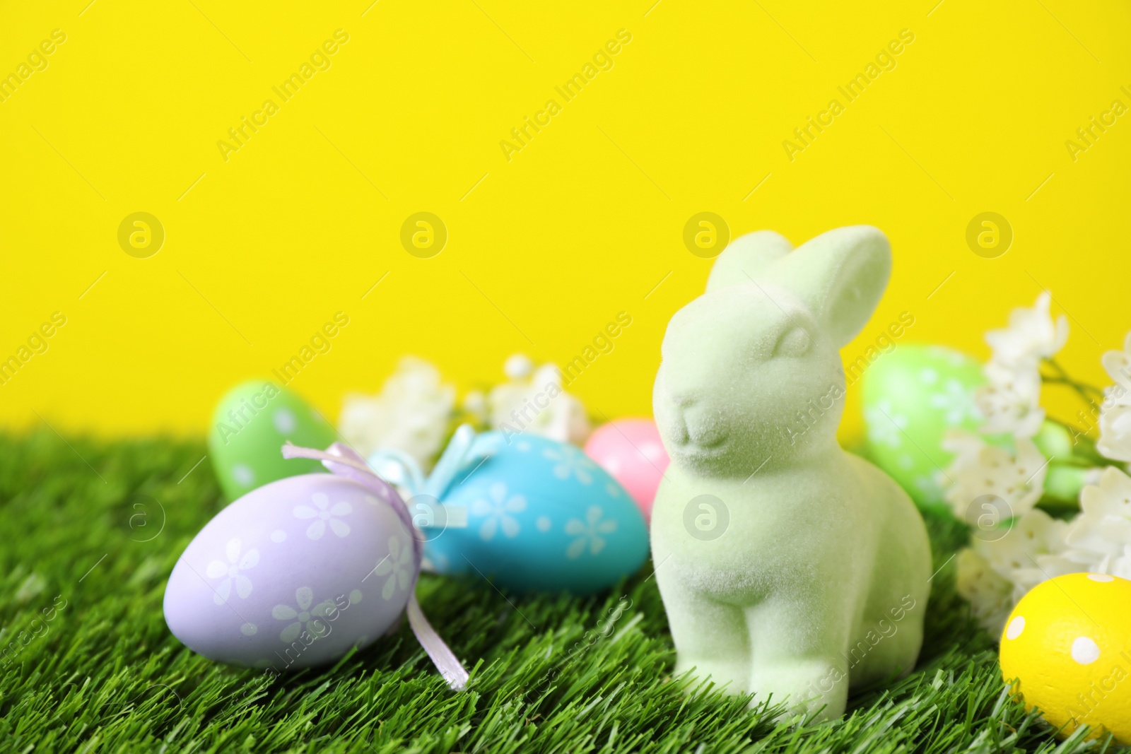 Photo of Easter bunny toy and dyed eggs on green grass against yellow background. Space for text