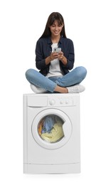 Photo of Beautiful woman using smartphone while sitting on washing machine with laundry against white background