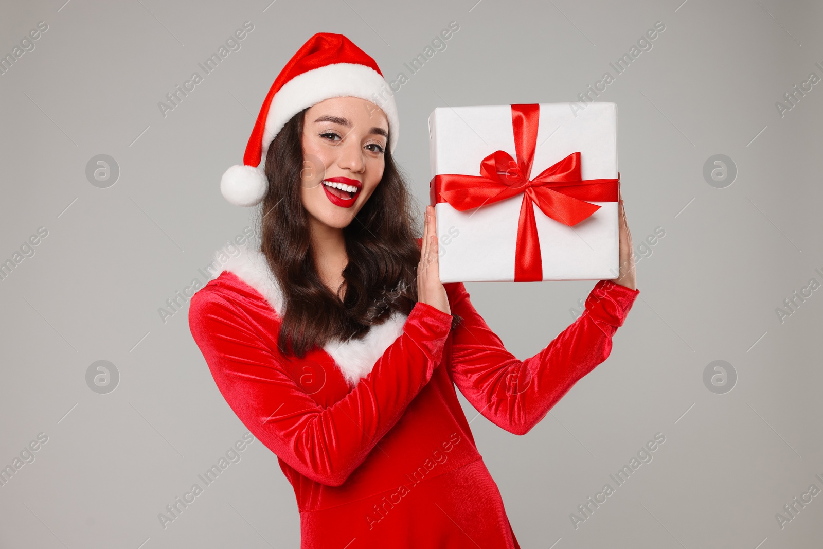 Photo of Beautiful young woman in red dress and Santa hat with Christmas gift on grey background