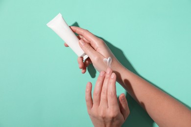 Photo of Woman with tube applying cosmetic cream onto her hand on turquoise background, top view