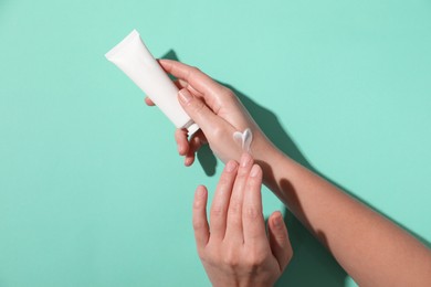 Woman with tube applying cosmetic cream onto her hand on turquoise background, top view