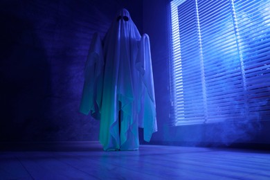 Creepy ghost. Woman covered with sheet near window in blue light, low angle view