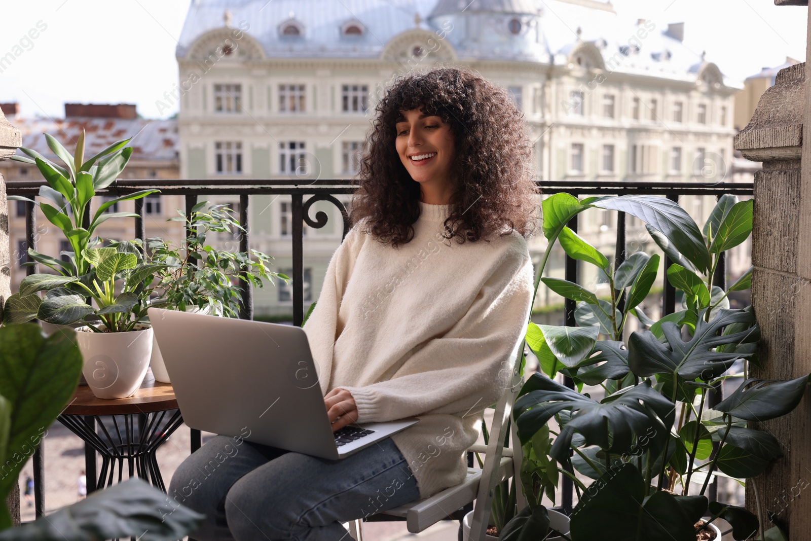 Photo of Beautiful young woman using laptop surrounded by green houseplants on balcony