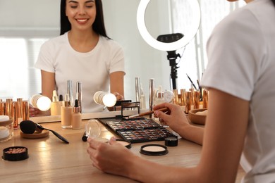 Photo of Beautiful young woman applying makeup at table with mirror and ring lamp