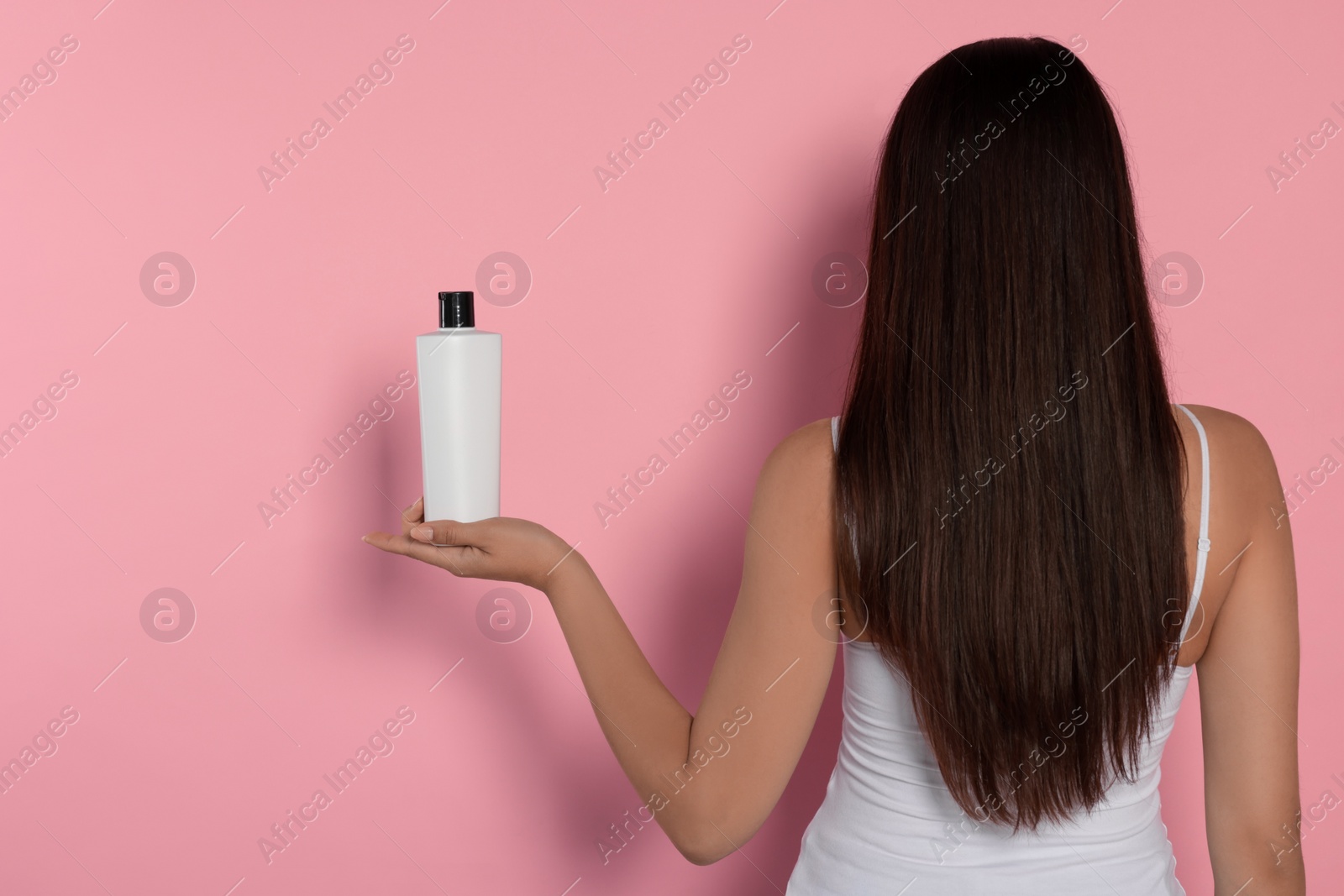 Photo of Young woman holding bottle of shampoo on pink background, back view. Space for text