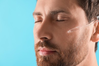 Handsome man applying cosmetic serum onto his face on light blue background, closeup