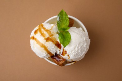 Photo of Scoops of tasty ice cream with caramel sauce, mint leaves and candies in paper cup on light brown background, top view