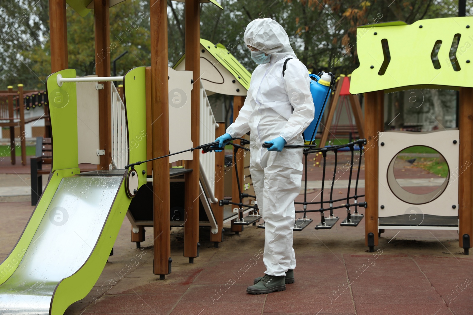 Photo of Woman wearing chemical protective suit with disinfectant sprayer on playground. Preventive measure during coronavirus pandemic