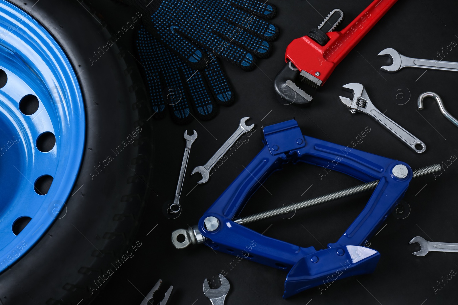 Photo of Car wheel, scissor jack, gloves and different tools on black background, flat lay