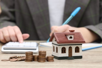 Photo of Woman writing at wooden table, focus on house model and stacked coins