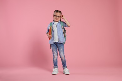 Happy schoolgirl in glasses with backpack and books on pink background