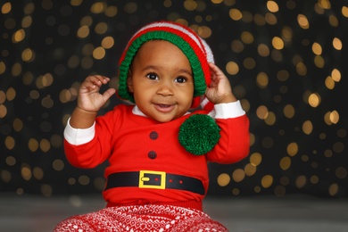 Image of Cute little African American baby wearing elf hat against blurred lights on dark background. Christmas celebration