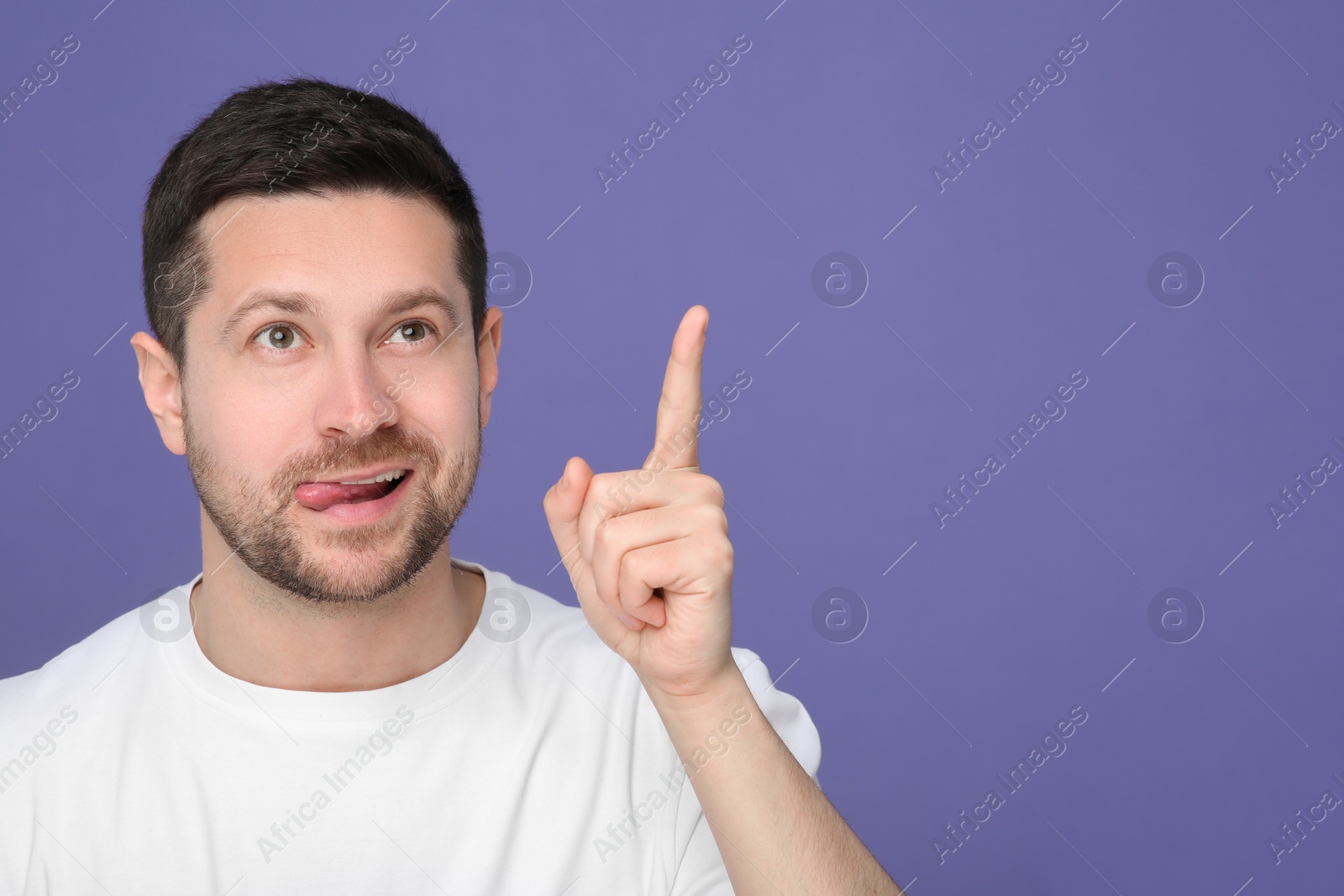 Photo of Man showing his tongue and pointing at something on purple background. Space for text