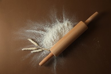 Photo of Flour, spikelets and rolling pin on brown table, top view