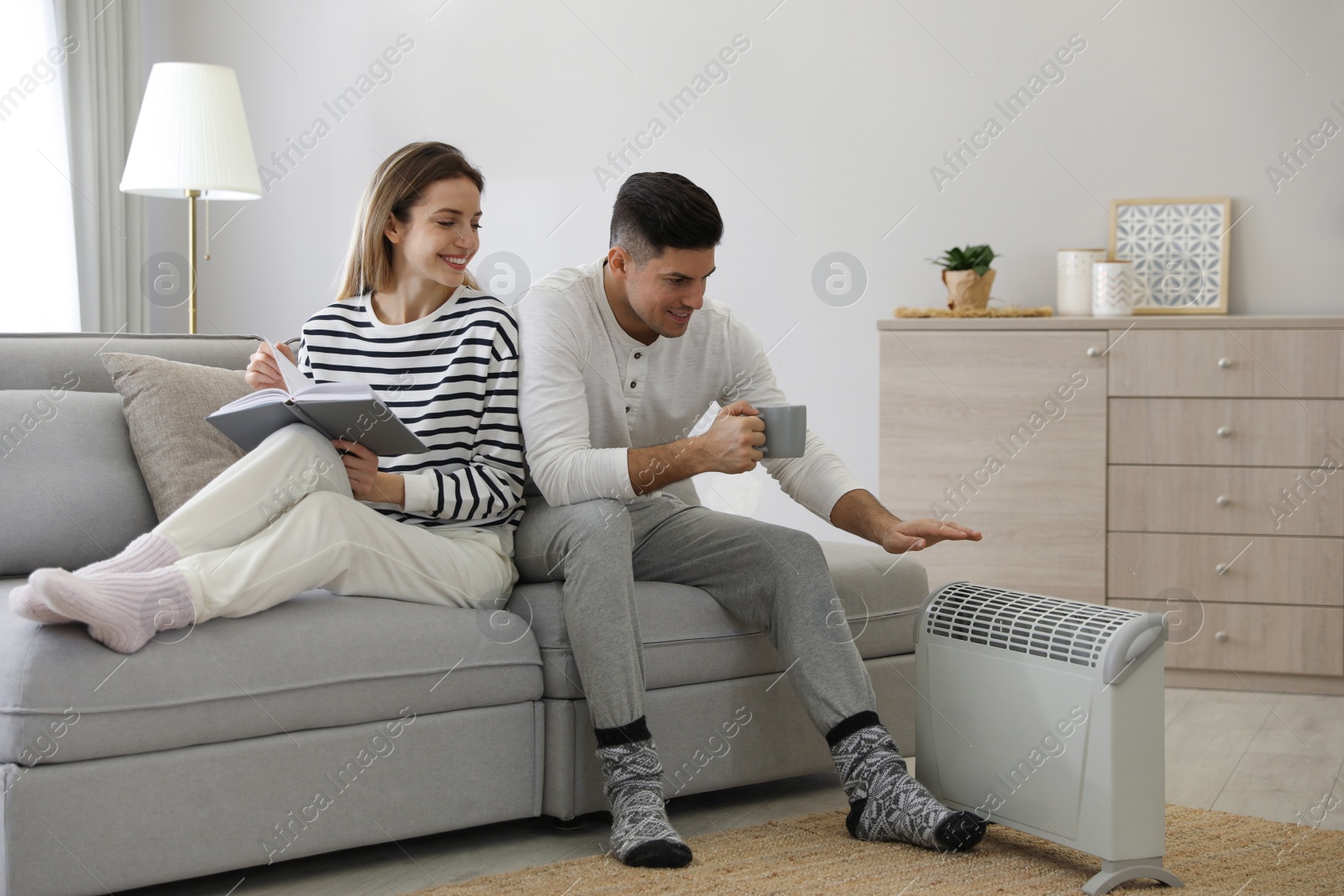 Photo of Happy couple sitting on sofa near electric heater at home