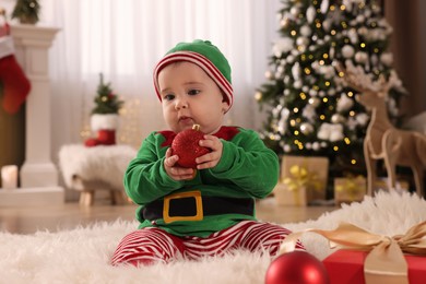 Photo of Baby in cute elf costume with Christmas ball on floor at home