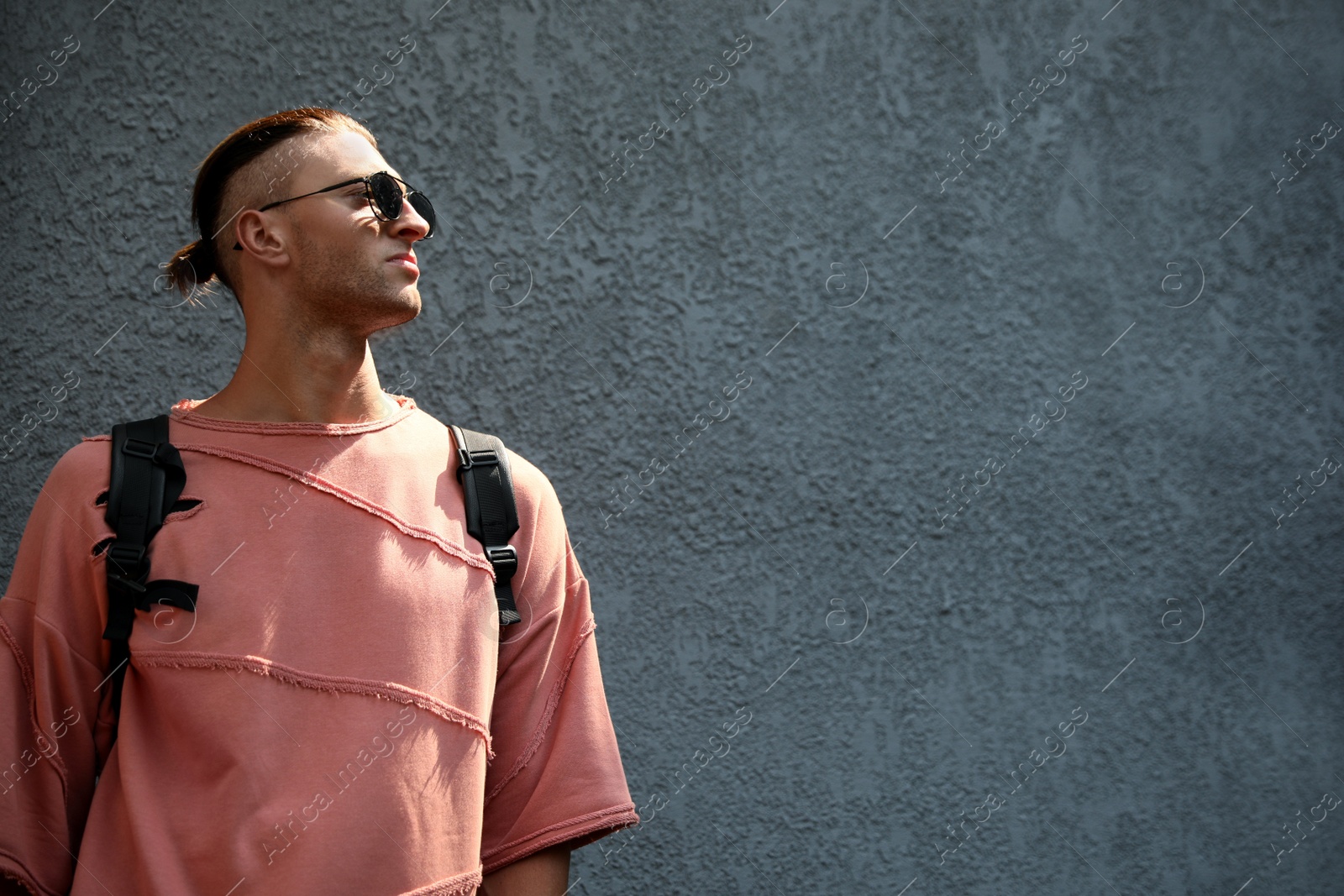 Photo of Handsome young man in stylish sunglasses and backpack near grey wall, space for text