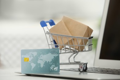 Photo of Online payment concept. Small shopping cart with bank card, boxes and laptop on table, closeup