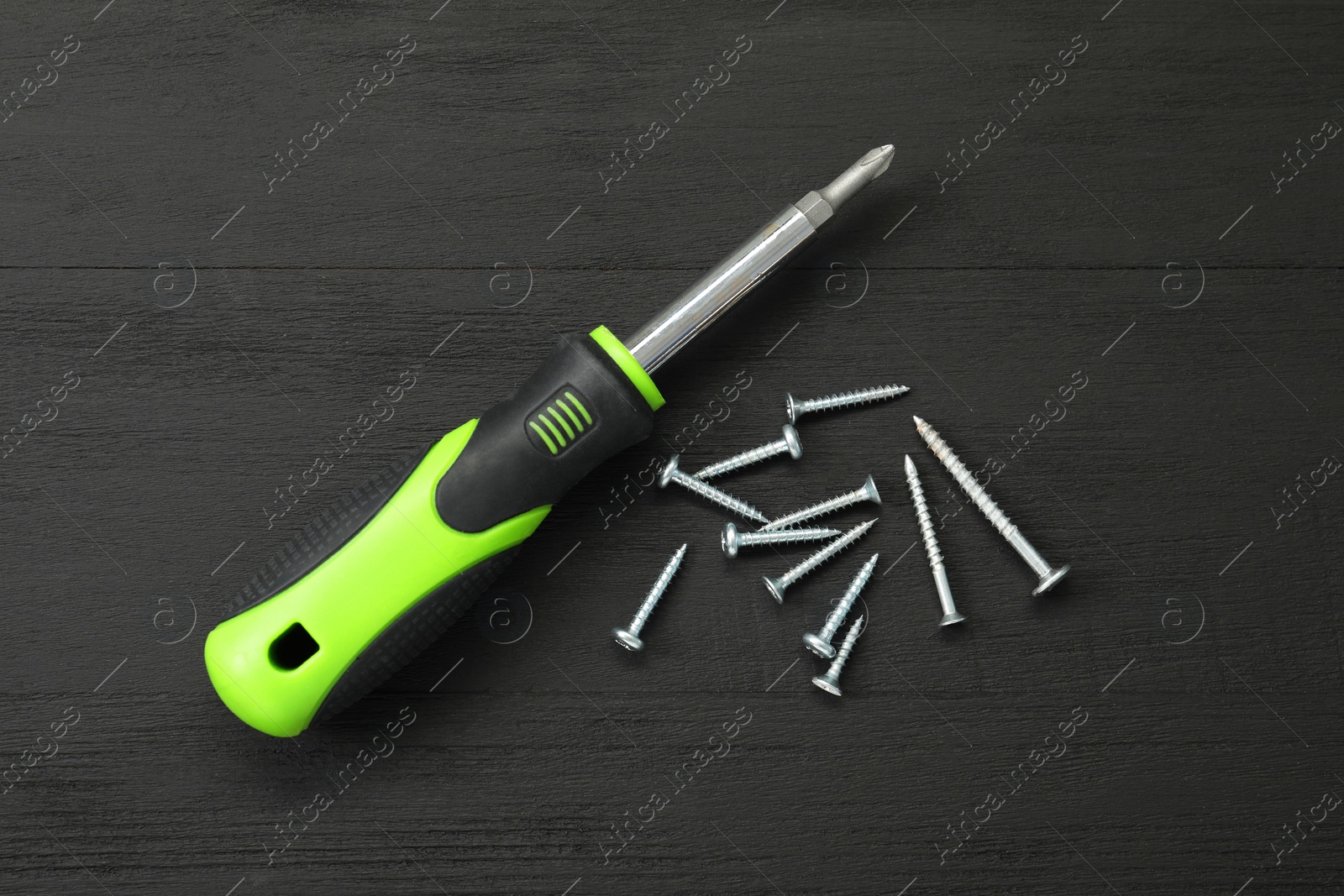 Photo of Screwdriver with green handle and screws on black wooden table, flat lay