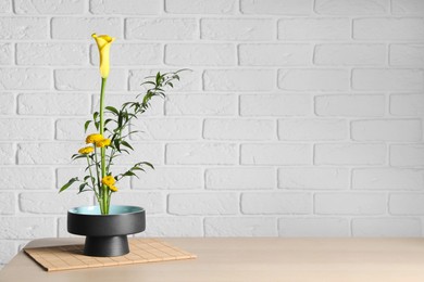 Photo of Beautiful ikebana for stylish house decor. Floral composition with fresh calla, chrysanthemum flowers and branches on wooden table near white brick wall, space for text