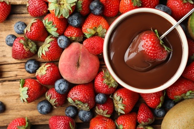 Photo of Fondue fork with strawberry in bowl of melted chocolate surrounded by other fruits on wooden table, flat lay