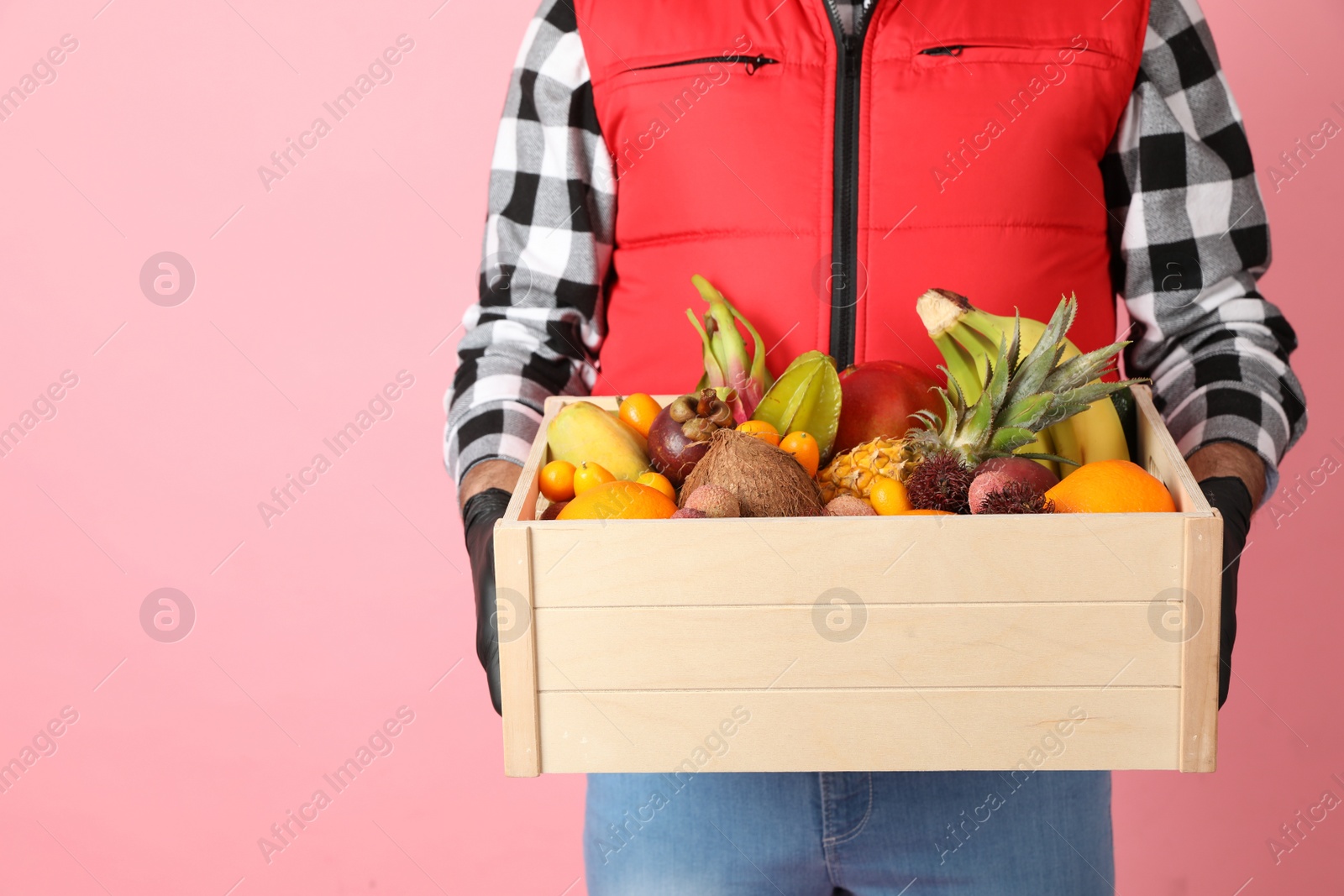 Photo of Courier holding crate with assortment of exotic fruits on pink background, closeup. Space for text