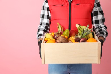 Photo of Courier holding crate with assortment of exotic fruits on pink background, closeup. Space for text
