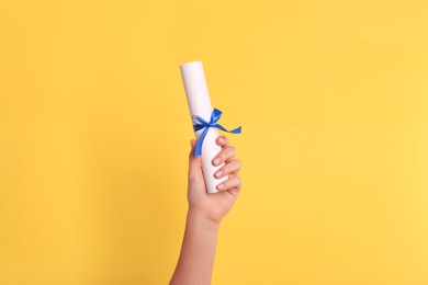 Photo of Student holding rolled diploma with blue ribbon on yellow background, closeup