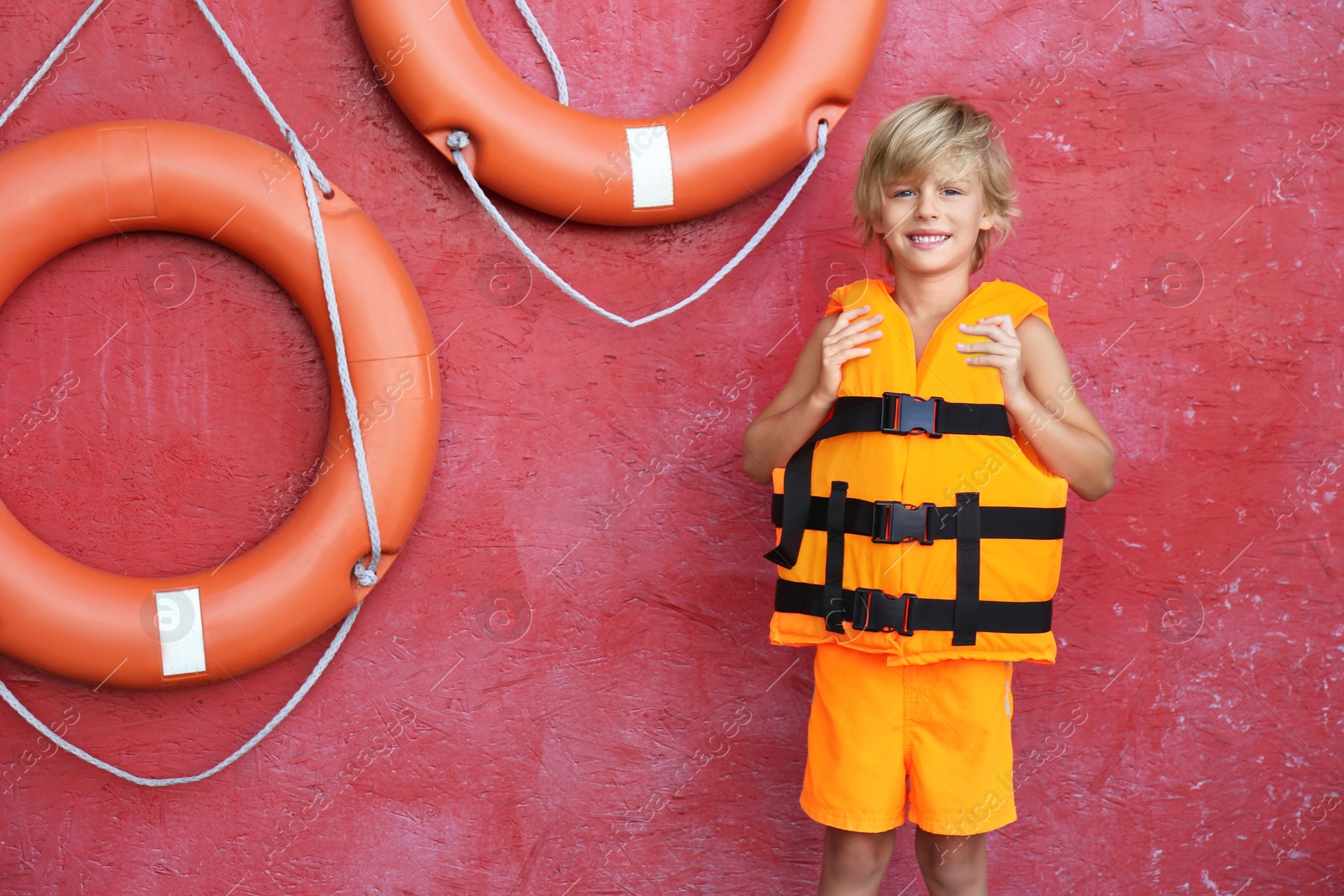 Photo of Little boy wearing orange life vest near red wall with safety rings