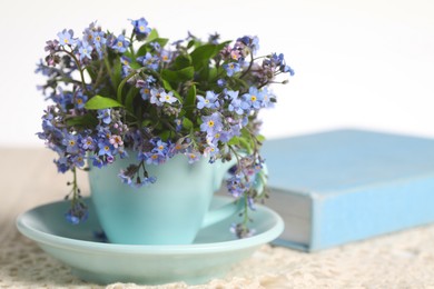 Photo of Beautiful forget-me-not flowers in cup, book and crochet tablecloth on table against white background, closeup