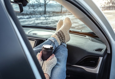 Photo of Young woman in warm socks holding her legs on car dashboard and drinking coffee. Cozy atmosphere