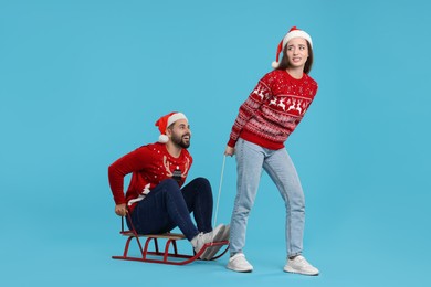 Photo of Young couple in Christmas sweaters. Woman pulling her man in sled on light blue background