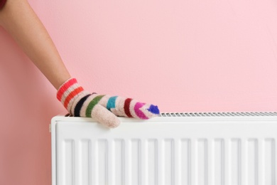 Photo of Woman in mitten warming hand on heating radiator near color wall. Space for text