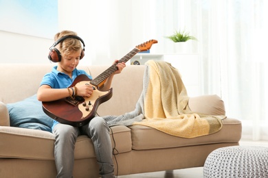 Photo of Cute little boy with headphones playing guitar on sofa in room. Space for text