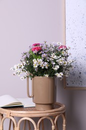 Photo of Stylish ceramic vase with beautiful flowers and open book on wicker table near light wall