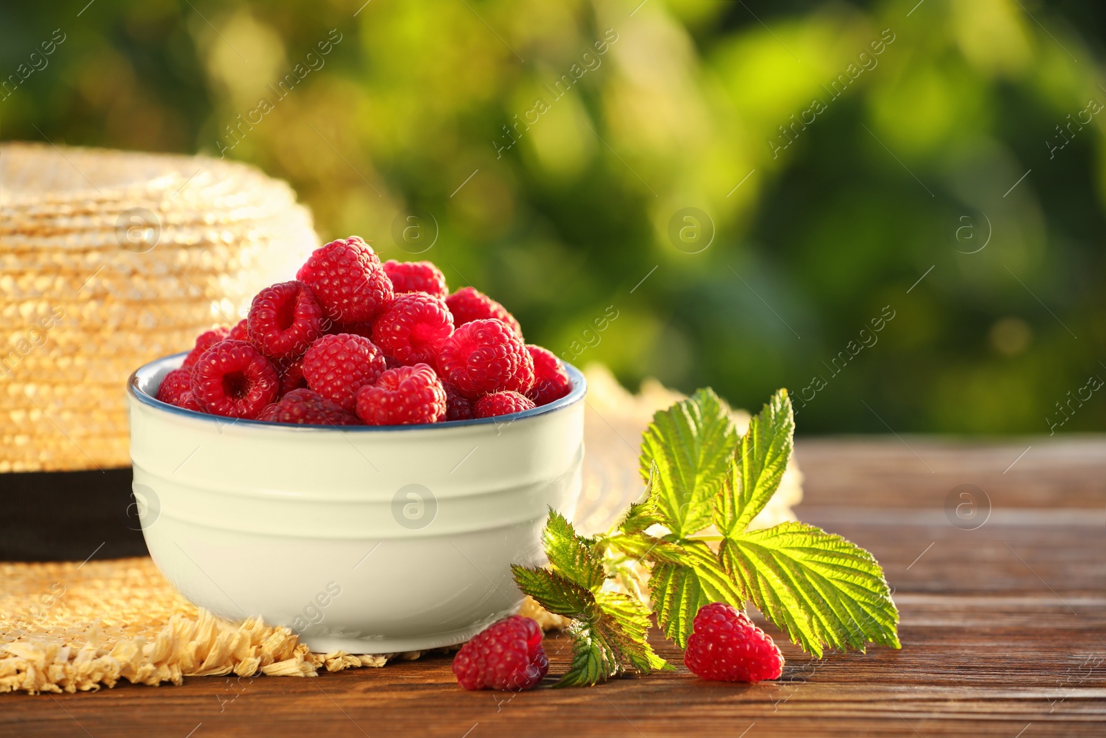 Photo of Tasty ripe raspberries, green leaves and straw hat on wooden table outdoors, closeup. Space for text