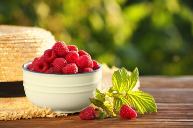 Photo of Tasty ripe raspberries, green leaves and straw hat on wooden table outdoors, closeup. Space for text