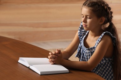 Photo of Cute little girl praying over Bible at table in room