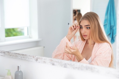 Photo of Young woman with eyelash loss problem looking in mirror indoors