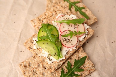 Photo of Tasty crispbreads with cream cheese, fresh cucumbers, radish and arugula on parchment paper, closeup