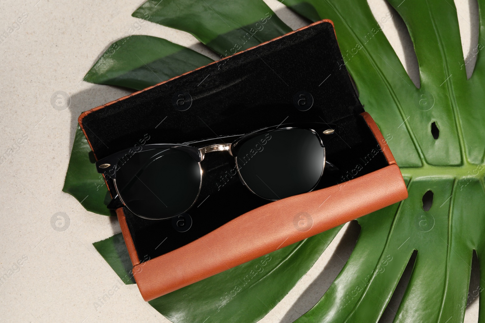 Photo of Stylish sunglasses in brown leather case and tropical leaf on sand, flat lay
