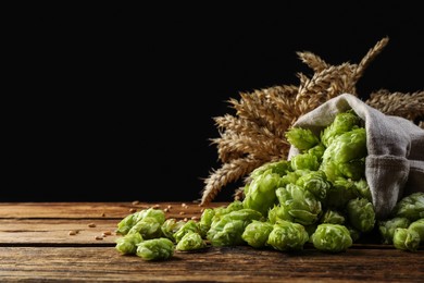 Photo of Overturned sack of hop flowers and wheat ears on wooden table against black background, space for text