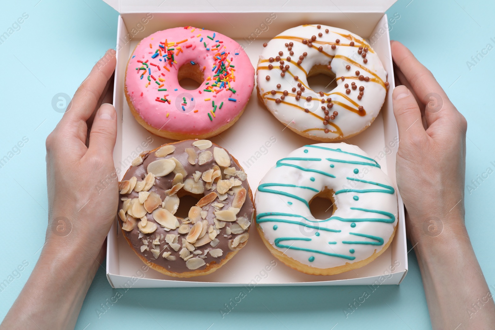 Photo of Woman holding box with tasty glazed donuts on light blue background, top view