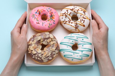 Woman holding box with tasty glazed donuts on light blue background, top view