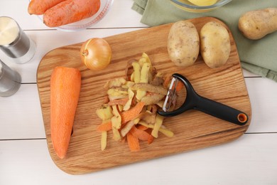 Peels of fresh vegetables and peeler on white wooden table, flat lay