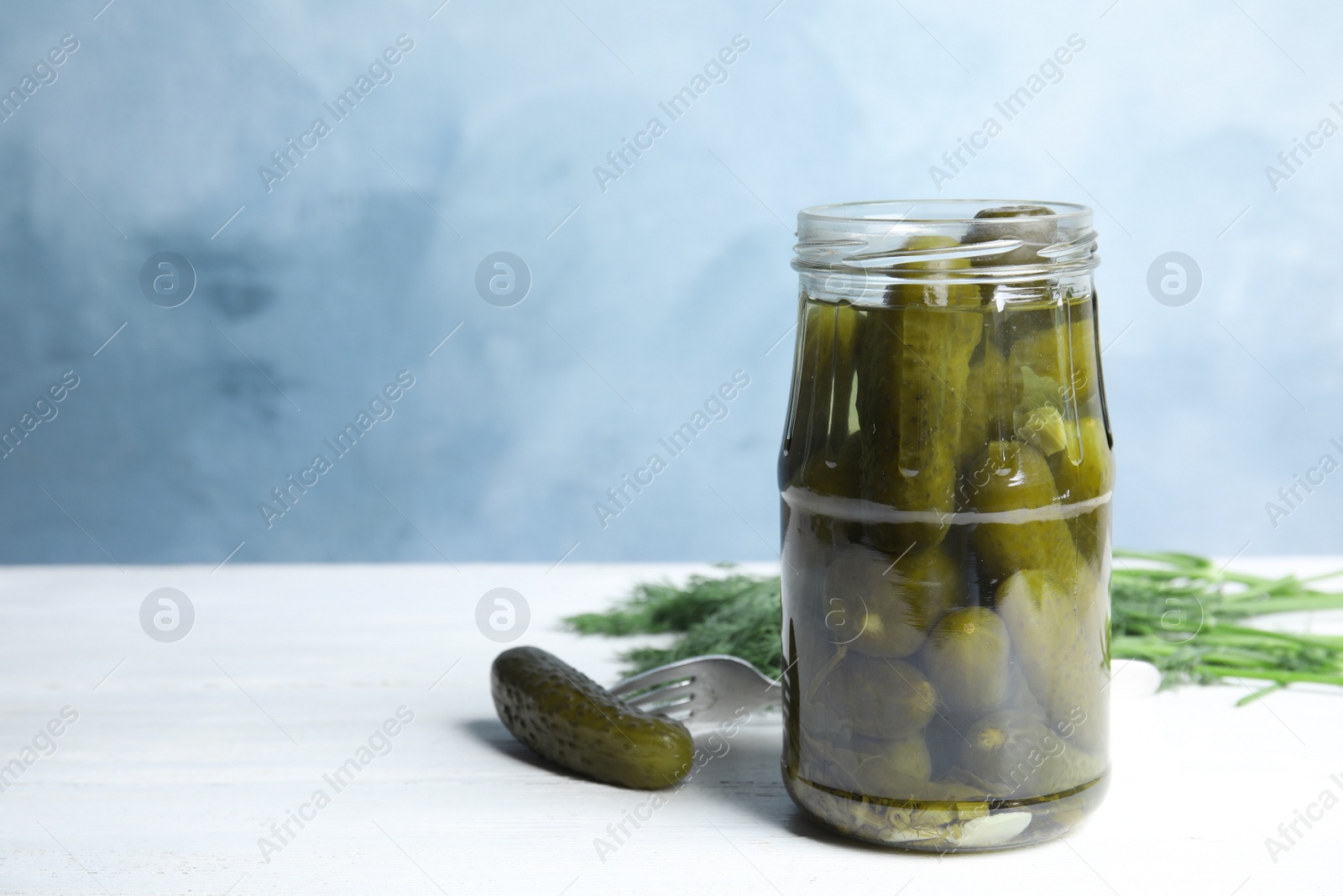 Photo of Jar with pickled cucumbers on white wooden table against blue background, space for text